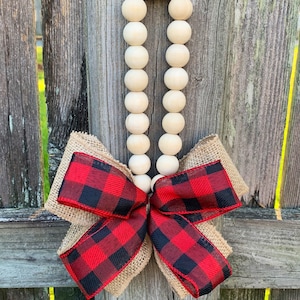 May include: A decorative wooden bead garland with a red and black buffalo plaid bow and burlap backing. The garland is hanging on a weathered wooden fence. The beads are light beige and round.