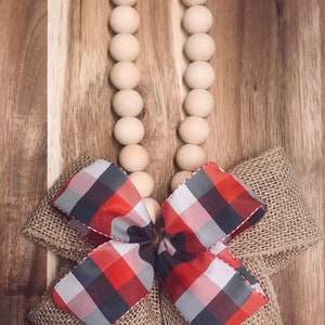 May include: A wooden bead garland with a red and black plaid bow tied with burlap. The garland is hanging on a wooden surface.