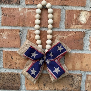 May include: A wooden bead garland with a burlap bow decorated with red, white, and blue stars. The garland is hanging on a brick wall.