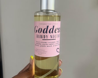 Può includere: Una bottiglia spray di vetro trasparente riempita con un liquido dorato, etichettata "Goddess Bawdy Nectar" con testo rosa e bianco. La bottiglia ha una pompa argentata ed è tenuta in mano con smalto giallo.