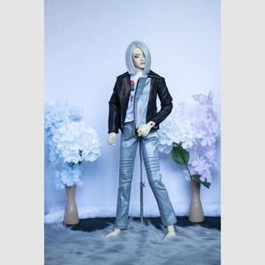 May include: A doll wearing a black leather jacket with a brown sheen, silver trousers, and a white t-shirt. The trousers have knee detailing. The doll has silver hair and is posed in front of white and blue floral arrangements.