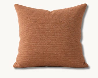 May include: A square throw pillow in a solid, earthy terracotta orange color. The fabric features a distinct, uniform woven texture with a visible grid-like pattern across the entire surface, creating a tactile and consistent appearance.