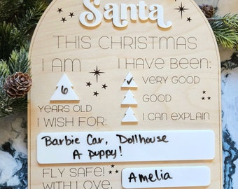 Peut inclure: Une pancarte en bois en forme d'arche avec les mots "Cher Père Noël" en haut. La pancarte comporte une liste de contrôle à remplir par un enfant, notamment son âge, son comportement et ses souhaits de Noël. L'enfant a écrit "Barbie Car, Dollhouse, A puppy!" pour ses souhaits. La pancarte comporte également un espace pour que l'enfant écrive son nom, et l'enfant a écrit "Amelia".