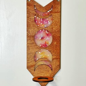May include: Wooden wall hanging with a crescent moon design and a candle holder. The moon shapes are filled with a pink and white resin with a floral pattern.