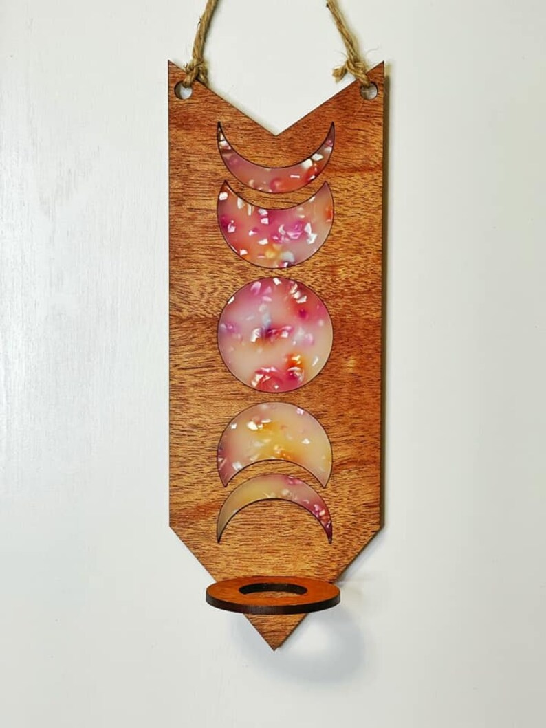 May include: Wooden wall hanging with a crescent moon design and a candle holder. The moon shapes are filled with a pink and white resin with a floral pattern.