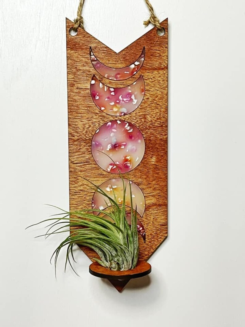May include: Wooden wall hanging with a moon phase design and a small plant holder. The moon phases are filled with colorful resin and the wood is stained a reddish brown.