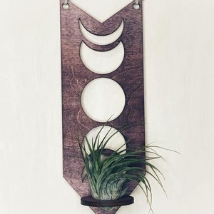 May include: A wooden wall hanging with a crescent moon design and a small shelf for a plant. The hanging is painted in a dark wood stain and has a rustic look.