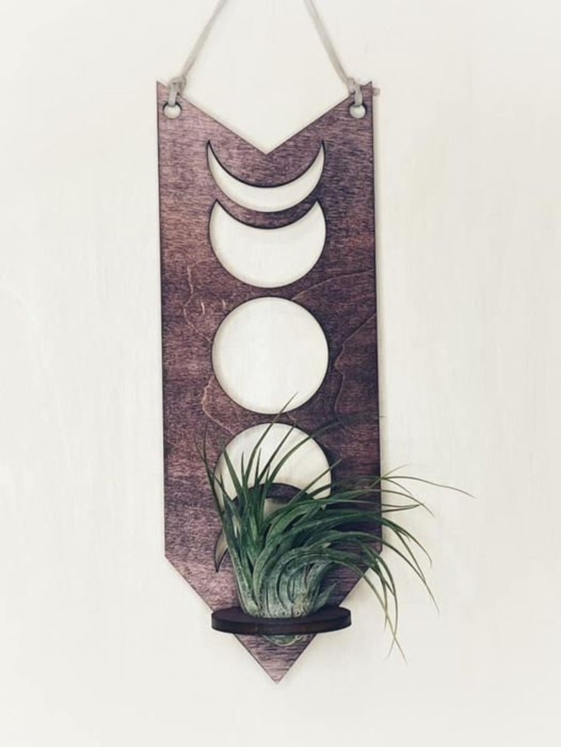 May include: A wooden wall hanging with a crescent moon design and a small shelf for a plant. The hanging is painted in a dark wood stain and has a rustic look.