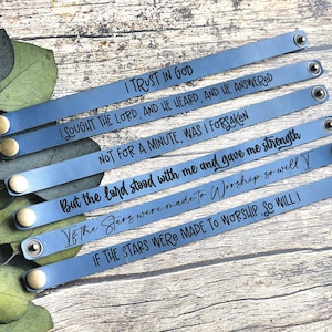 May include: Six blue leather bracelets with inspirational quotes in black lettering. Each bracelet has a snap closure with a brass-coloured button. The quotes include phrases like "I trust in God" and "If the stars were made to worship, so will I."