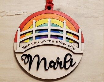 Peut inclure: Une décoration en bois en forme de pont arc-en-ciel avec le texte "See you on the other side" et le nom "Marli" écrit en dessous. La décoration est décorée avec un arc-en-ciel de couleurs.