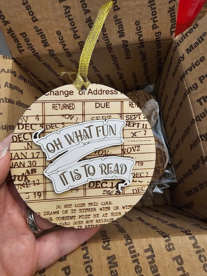 Peut inclure: Une d&eacute;coration en bois en forme de carte de changement d'adresse vintage. La d&eacute;coration pr&eacute;sente le texte "Oh What Fun It Is To Read" dans une banni&egrave;re.