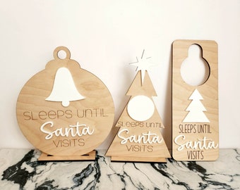 Peut inclure: Trois pancartes en bois avec le texte "Sleeps Until Santa Visits" en lettres blanches. Les pancartes sont en forme de cloche, de sapin de Noël et de porte-manteau.
