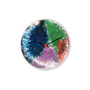 Puede incluir: Un reloj redondo con fondo blanco y arte abstracto colorido en tonos de azul, verde, morado y rojo. Las manecillas del reloj son negras con una punta roja.