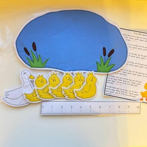 May include: A felt play set for the children's book "Five Little Ducks" featuring a pond, five yellow ducklings, a mother duck, and a number line from 0 to 10. The text from the book is printed on a separate piece of felt.