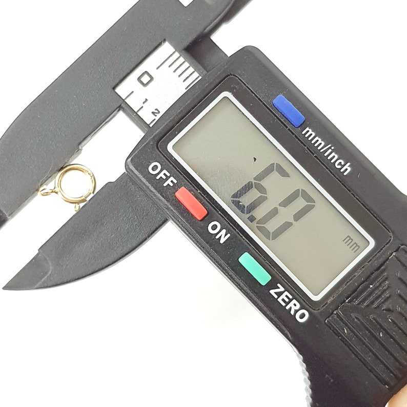 May include: A black digital caliper measuring a gold jump ring. The display reads 6.0 mm.