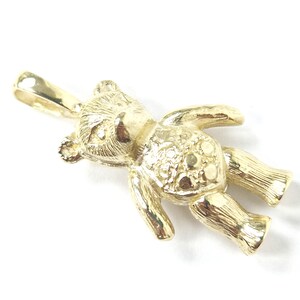 May include: A gold-colored teddy bear pendant with a textured surface. The bear is lying down with its arms and legs outstretched. The pendant has a loop at the top for a chain.