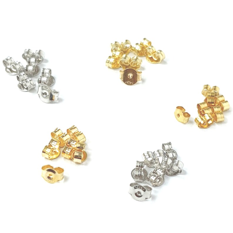 May include: A close-up of earring backs in three different colors: silver, gold, and rose gold. The earring backs are made of metal and have a small hole in the center. They are designed to be used with post earrings.