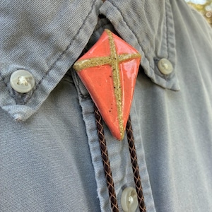 May include: A coral-colored bolo tie with a kite-shaped pendant. The pendant features a gold cross design and is suspended from a brown braided leather cord. The bolo tie is worn over a light blue button-down shirt.