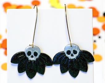 Puede incluir: Un par de pendientes colgantes con un diseño de calavera plateada sobre una base en forma de pétalo verde oscuro. Los pendientes tienen ganchos largos y finos de color dorado. El fondo es blanco con confeti naranja y amarillo disperso.