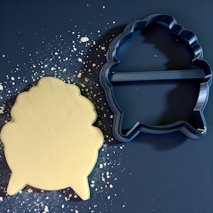 Puede incluir: Un cortador de galletas en forma de caldero azul oscuro junto a una galleta horneada con la misma forma. La galleta es de color beige claro y el cortador es de un azul más oscuro. Se espolvorea polvo blanco alrededor de la galleta y el cortador sobre una superficie azul oscuro.