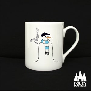 May include: White ceramic mug with a black and white illustration of a snowman wearing a blue and white scarf in front of a building with a tall antenna. The mug has a black handle.