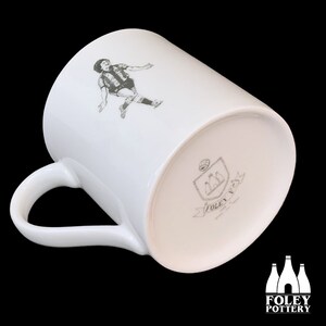 May include: White ceramic mug with a black and white illustration of a football player on one side and a black and white logo on the other side. The logo features three bottles and the text "FOLEY POTTERY".