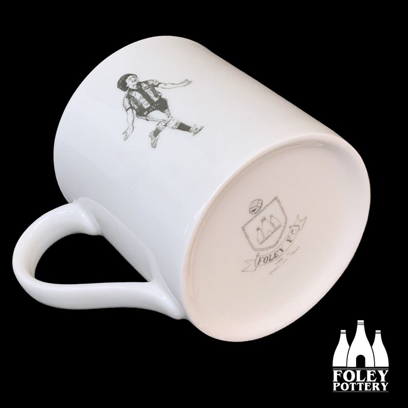 May include: White ceramic mug with a black and white illustration of a football player on one side and a black and white logo on the other side. The logo features three bottles and the text "FOLEY POTTERY".