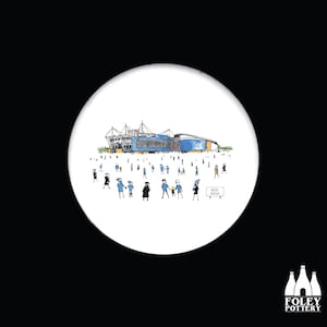 May include: A white ceramic coaster with a cartoon illustration of a stadium with people in front of it. The stadium is blue and white with the word "Etihad" on the side. The sign in front of the stadium says "Etihad Arena".