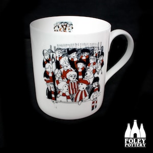 May include: White ceramic mug with a black and white illustration of a crowd of people wearing red and white scarves. The mug has a black handle and the words "FOLEY POTTERY" are printed on the bottom.