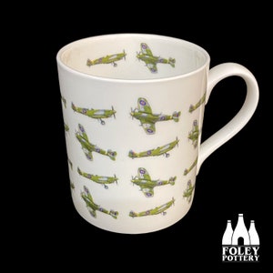 May include: White ceramic mug with a repeating pattern of green and brown fighter planes. The mug has a handle and the words "FOLEY POTTERY" are printed on the bottom.