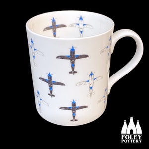 May include: White ceramic mug with a repeating pattern of blue and gray airplanes. The mug has a handle and the words "FOLEY POTTERY" are printed on the bottom.