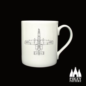 May include: A white ceramic mug featuring a black line drawing of an A-10 Thunderbolt II aircraft on the front. The mug has a handle on the right side.