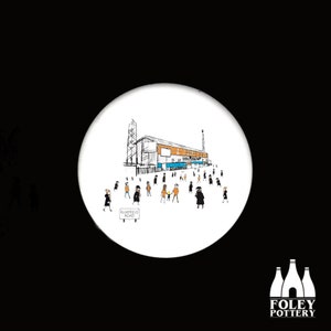 May include: A white ceramic coaster with a black and white illustration of a stadium with people walking towards it. The stadium is labeled "Bloomfield Road".