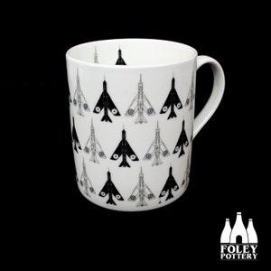 May include: A white ceramic mug with a black and white pattern of fighter jets. The mug has a black handle and the words "FOLEY POTTERY" are printed on the bottom.