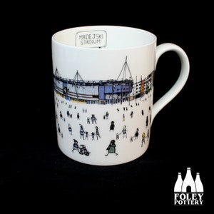 May include: White ceramic mug with a black and white illustration of the Madejski Stadium in Reading, England. The stadium is depicted with a crowd of stick figures in front of it. The text "Madejski Stadium" is printed on the mug.