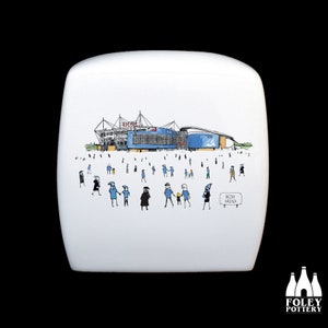 May include: A white ceramic box with a hand-drawn illustration of a stadium with people in front of it. The stadium is blue and white with the word "Etihad" on the side. The text "Etihad Arena" is on a sign in the bottom right corner of the image.