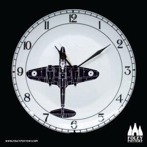 May include: A white ceramic clock face with black numbers and hands. A black silhouette of an airplane is in the center of the clock face.