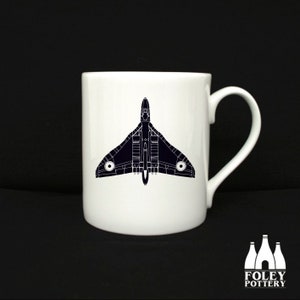 May include: A white ceramic mug with a black silhouette of a jet plane on the front. The mug has a handle and the text "FOLEY POTTERY" is printed on the bottom.
