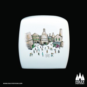 May include: A white ceramic toilet tank lid with a hand-painted illustration of a pottery factory. The illustration features a group of people walking around the factory, which has several tall chimneys. The text "LANGSTONE POTTERY" is visible on a sign in the illustration.