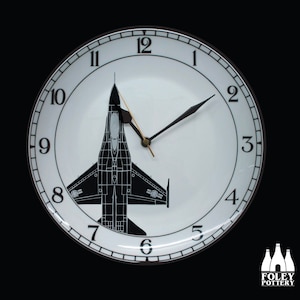 Könnte beinhalten: Eine weiße Keramik-Uhr mit schwarzen Zahlen und Zeigern. Im Zentrum des Zifferblatts befindet sich eine schwarze Silhouette eines Kampfjets.