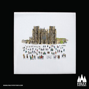 Puede incluir: Un azulejo de cerámica blanco con una ilustración dibujada a mano de la Catedral de Wells en Inglaterra. La catedral está representada en marrón, gris y negro, con una multitud de personas de varios colores de pie frente a ella. Un cartel dice "Wells Cathedral".