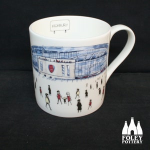 Puede incluir: Taza de cerámica blanca con una ilustración de dibujos animados de un estadio, personas y el texto "Highbury" en el lateral.