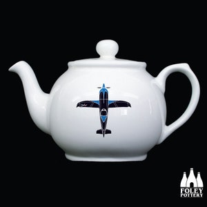 May include: White ceramic teapot with a black and blue airplane design on the front. The teapot has a round body and a spout on the left side.