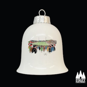 May include: White bell-shaped Christmas ornament with a silver cap and a loop for hanging. The ornament features a watercolor illustration of a stadium filled with people. The Foley Pottery logo is at the bottom.