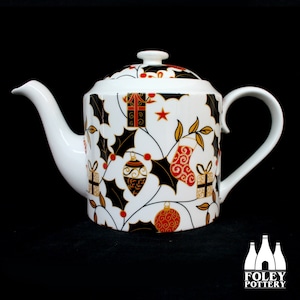 Puede incluir: Tetera de cerámica blanca con un diseño navideño negro y dorado. La tetera presenta hojas de acebo, adornos y cajas de regalo. La tetera está hecha por Foley Pottery.