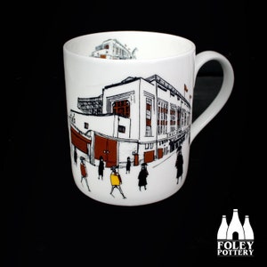 Puede incluir: Taza de cerámica blanca con una ilustración en blanco y negro de un estadio y personas caminando frente a él. El estadio es marrón con una puerta roja. La taza tiene un asa negra.