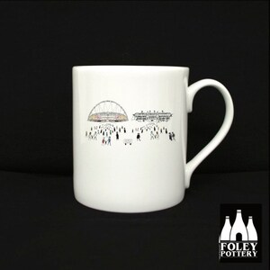 May include: White ceramic mug with a black and white illustration of a stadium and people walking towards it. The mug has a black handle and the words "FOLEY POTTERY" are printed on the bottom.