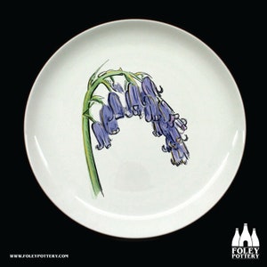May include: White ceramic plate featuring a hand-painted illustration of bluebell flowers. The flowers are depicted in shades of purple and blue with green stems. The plate has a simple, elegant design, suitable for serving food or as a decorative piece.