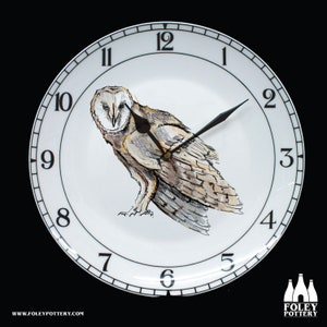 May include: A white ceramic clock with black Roman numeral hour markers and black hands. The clock face features a hand-painted barn owl with brown and white feathers. The clock is 12 inches in diameter and is made by Foley Pottery.