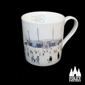May include: White ceramic mug with a black and white illustration of the City of Manchester Stadium. The mug has the text "City of Manchester Stadium" on the side.